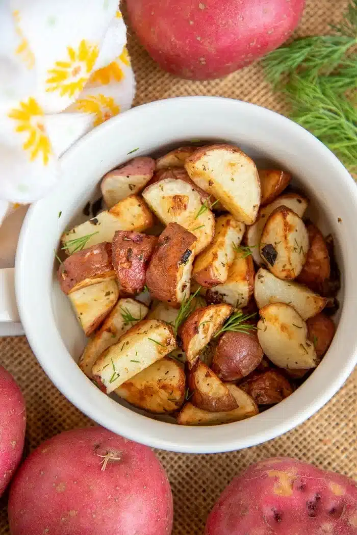 A white bowl filled with perfectly roasted air fryer red potatoes, garnished with herbs, sits on a burlap surface surrounded by whole red potatoes and a sprig of dill. A white cloth adorned with yellow flowers peeks from the corner, adding a touch of charm.