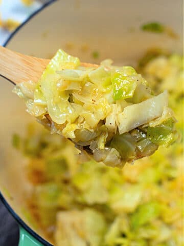 A wooden spoon holds a portion of stewed cabbage with visible seasonings. The cabbage is nestled in a green pot, and the background showcases more of this hearty, flavorful dish.