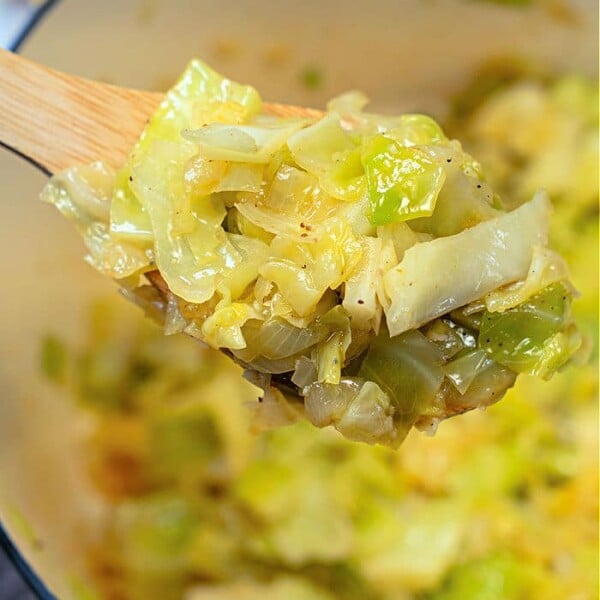 A wooden spoon holds a portion of stewed cabbage with visible seasonings. The cabbage is nestled in a green pot, and the background showcases more of this hearty, flavorful dish.