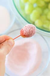 A hand holding a grape coated in pink sugary powder, showcasing how to make candied grapes with Jello; green grapes are visible in a bowl in the background.