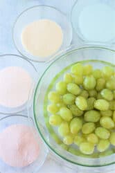 A bowl of green grapes, waiting to be transformed into candied delights with Jello, is surrounded by bowls of white, pink, peach, and cream-colored powders on a marble surface.