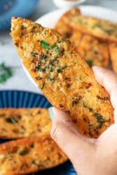 A hand holding a slice of golden garlic bread, freshly made from scratch and garnished with herbs, with more slices on a blue plate in the background.