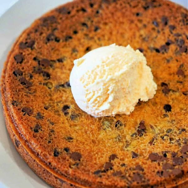 A large pizookie, crisped to perfection in an air fryer, sits on a white plate. It's topped with a scoop of vanilla ice cream melting into the slightly browned cookie, with chocolate chips delightfully scattered throughout.