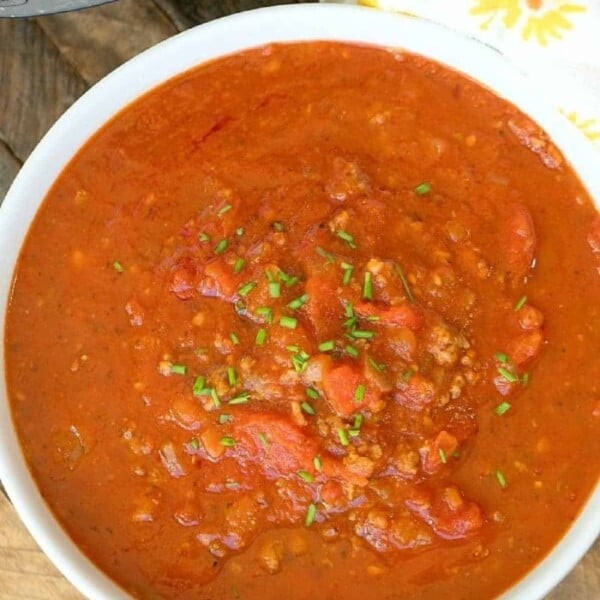 A bowl of homemade spaghetti sauce garnished with chopped herbs sits on a wooden table near a patterned yellow and white towel, while an instant pot spaghetti sauce simmers nearby, enhancing the cozy kitchen ambiance.