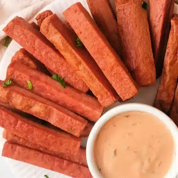 A plate of rectangular fried Spam fries is arranged around a small dish of creamy dipping sauce on a white plate. Some parsley leaves are scattered for garnish. The background is a light-colored fabric.