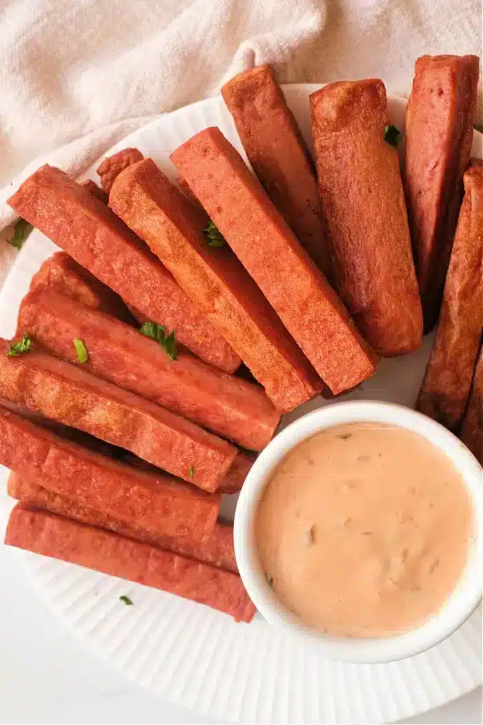 A plate of rectangular fried Spam fries is arranged around a small dish of creamy dipping sauce on a white plate. Some parsley leaves are scattered for garnish. The background is a light-colored fabric.