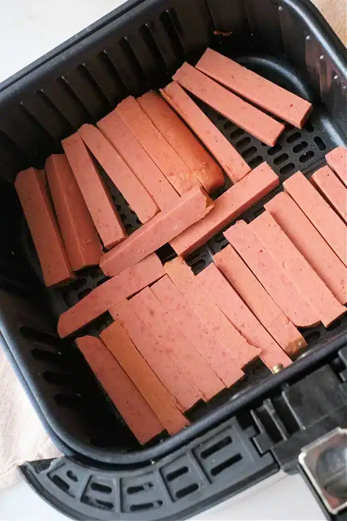 Sliced pieces of spam are arranged in the basket of an air fryer, ready for cooking