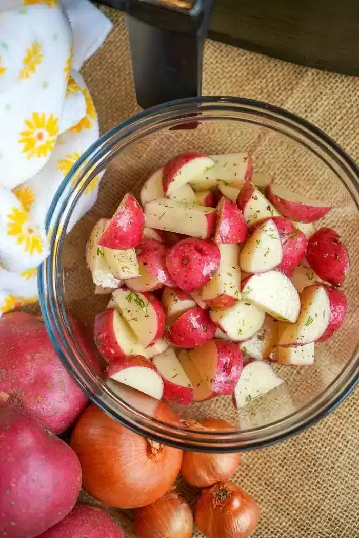 A glass bowl filled with chopped red potatoes seasoned with herbs is placed on a burlap surface, ready to become delicious air fryer red potatoes. Surrounding the bowl are whole red potatoes, onions, and a white cloth with yellow floral patterns.