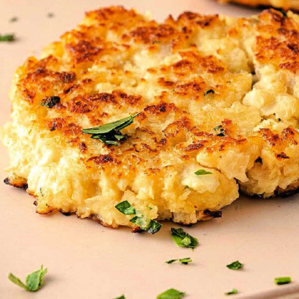 Close-up of a golden-brown, crispy cauliflower pancake garnished with chopped parsley. The pancake boasts a slightly uneven surface, revealing its texture. Additional pancakes are partially visible on a light-colored surface.