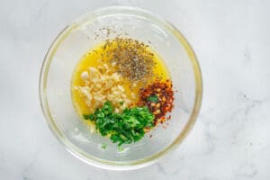 A glass bowl with melted butter, chopped garlic, parsley, red pepper flakes, and Italian herbs on a white surface—just what you need for learning How to Make Garlic Bread From Scratch.