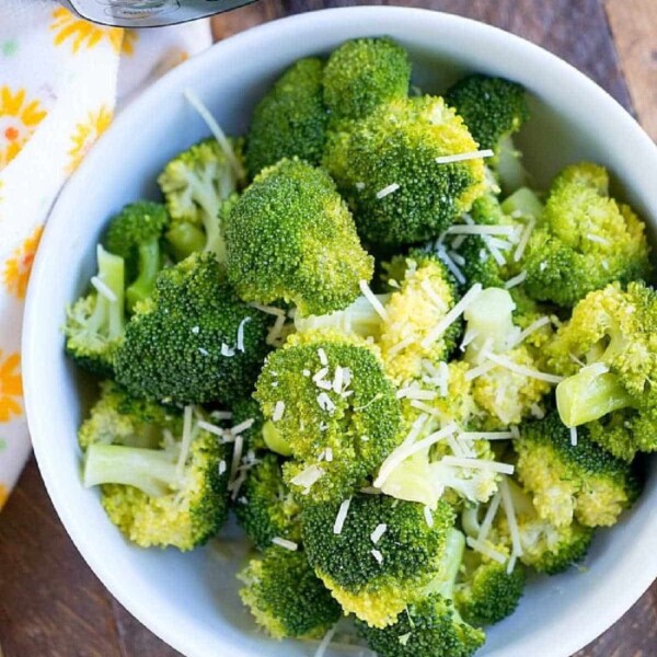 A bowl filled with freshly cooked broccoli florets, sprinkled with grated cheese, makes a perfect instant pot side dish. Positioned on a wooden surface next to your trusty Instant Pot pressure cooker, it sits elegantly beside a white cloth adorned with yellow flowers.