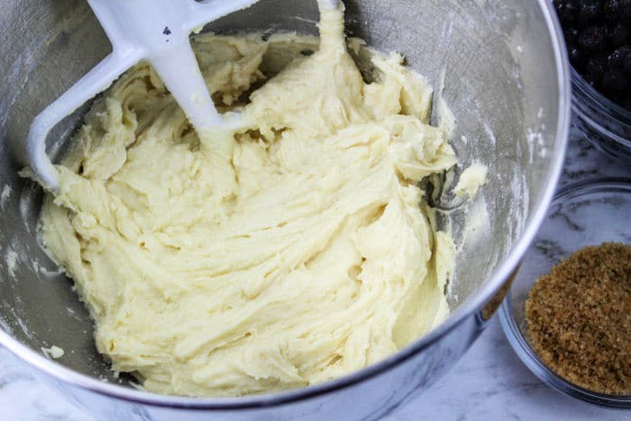 muffin batter is being mixed in a metal bowl with a paddle attachment, surrounded by bowls of brown sugar and chocolate chips. The aroma of jumbo blueberry muffins from the oven adds to the cozy kitchen ambiance.