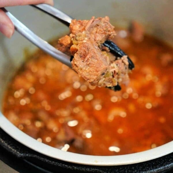 A close-up shows a hand holding metal tongs with a piece of succulent carnitas above a pot brimming with meat in a rich, red sauce. The pot appears to be part of a pressure cooker, ensuring the flavors meld perfectly.