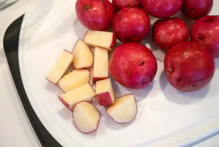 Red potatoes are on a cutting board, ready for the air fryer; some are whole while others are cut into chunks. They boast smooth, reddish skin and pale yellow flesh. The white cutting board with a black handle complements this vibrant preparation perfectly.