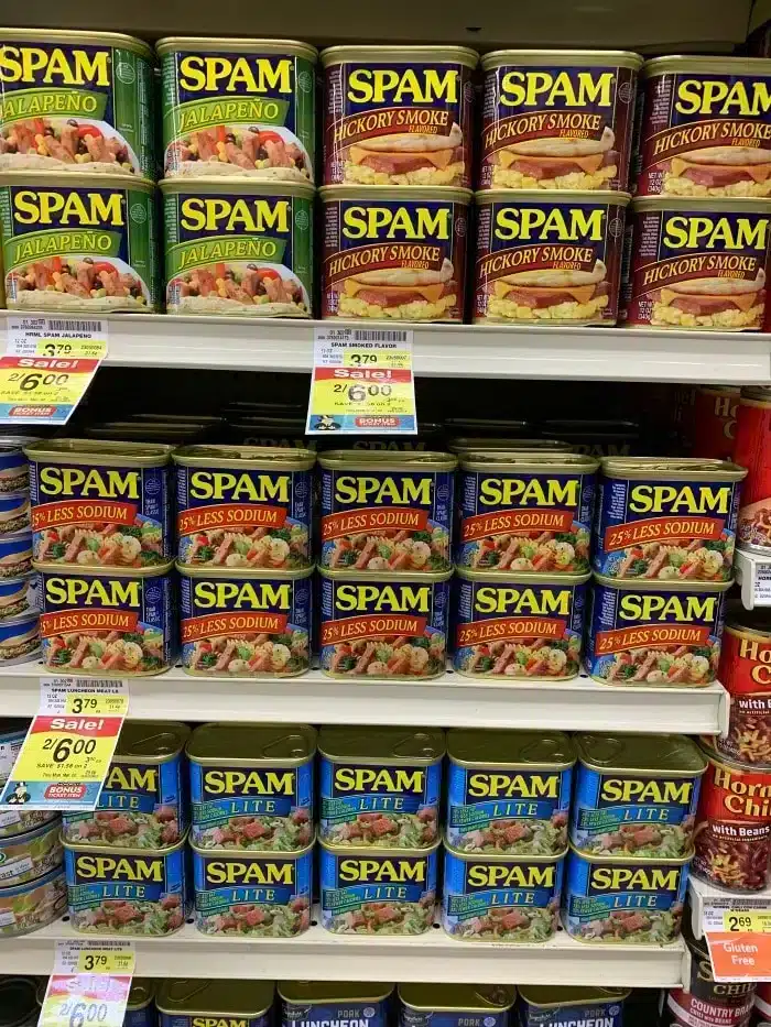 Shelves stocked with various flavors of Spam, including Spam Fries, Jalapeño, Hickory Smoke, 25% Less Sodium, and Lite. Price tags and promotions are visible beneath the cans.