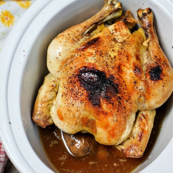 A frozen whole chicken rests, perfectly seasoned with pepper, in a Crockpot. It's nestled in a pool of savory juices, while a hint of yellow floral cloth adds a touch of charm to the scene.