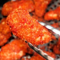 Tongs hold a piece of breaded and fried chicken, seasoned to perfection with Doritos Ranch Dipped Hot Wings spices, surrounded by more tantalizing pieces in the background.