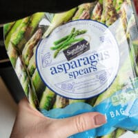 A hand holds a package of Signature Select frozen asparagus spears, perfect for the air fryer. The front label features images of asparagus and highlights product features such as being steamable and resealable.