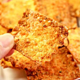 A close-up of a hand holding a square, crispy cheese cracker with jagged edges. Several other crackers are visible in the background on a plate, resembling delightful pasta chips with their golden-brown hue and textured surface.