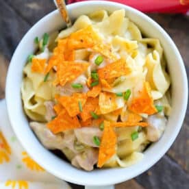 A bowl of creamy pasta reminiscent of a pressure cooker Dorito casserole, topped with crushed Doritos and chopped chives, sits beside a spoon and a Doritos bag.