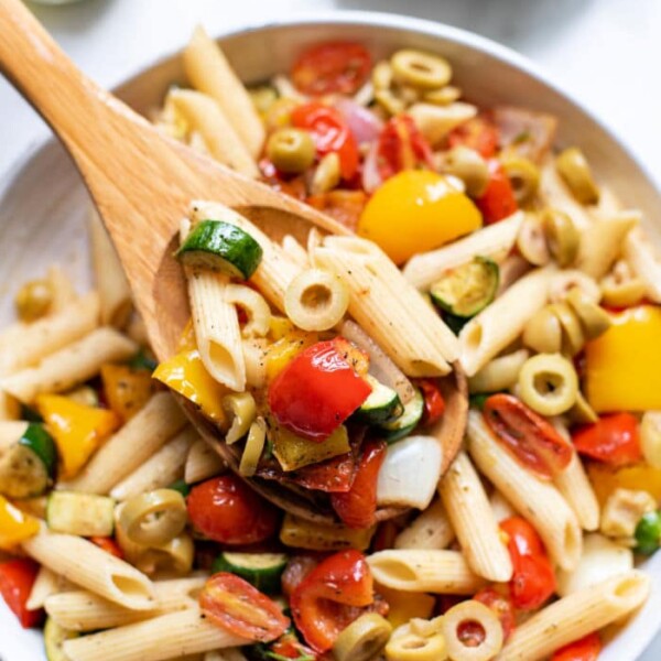 A bowl of roasted vegetable pasta salad with penne, tomatoes, olives, zucchini, and bell peppers, served with a wooden spoon.