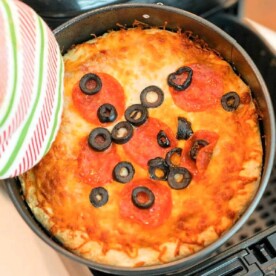 A baked air fryer pizza with a golden cheese crust, topped with sliced pepperoni and black olives, is being held in a pan using a striped oven mitt. The edges are slightly browned, suggesting it's freshly cooked.