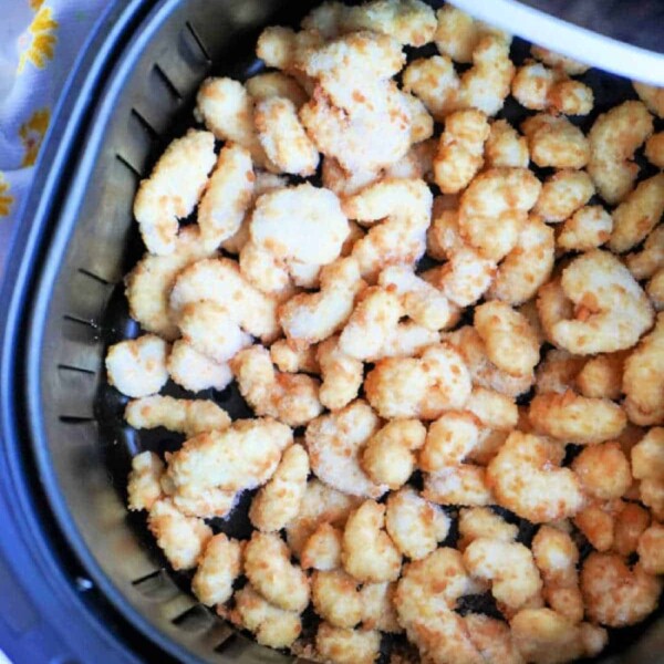 An air fryer basket brimming with crispy, golden-brown popcorn chicken pieces sits enticingly on a surface, alongside a yellow floral-patterned cloth. It's the perfect setup to savor frozen popcorn shrimp in the air fryer next time.