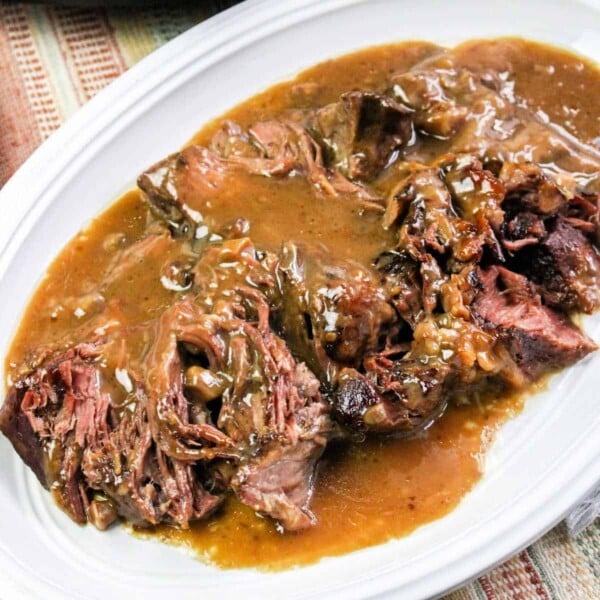 Tender pot roast, cooked to perfection in a pressure cooker, is smothered in gravy on a white plate, served on a striped tablecloth.