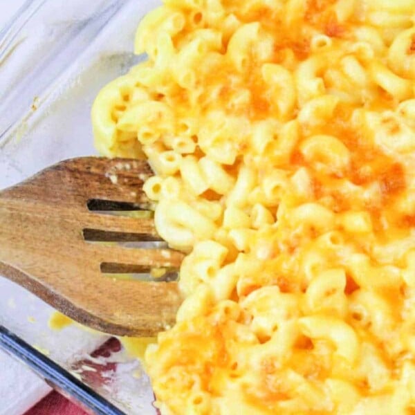 Baked mac and cheese, reminiscent of a Copycat Chick Fil A Mac and Cheese, sits invitingly in a glass dish with a wooden spatula on a red and white cloth.