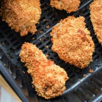 Four pieces of crispy, golden-brown gluten-free chicken are arranged in an air fryer basket. The chicken appears evenly cooked and crunchy. The basket rests on a light-colored wooden surface.