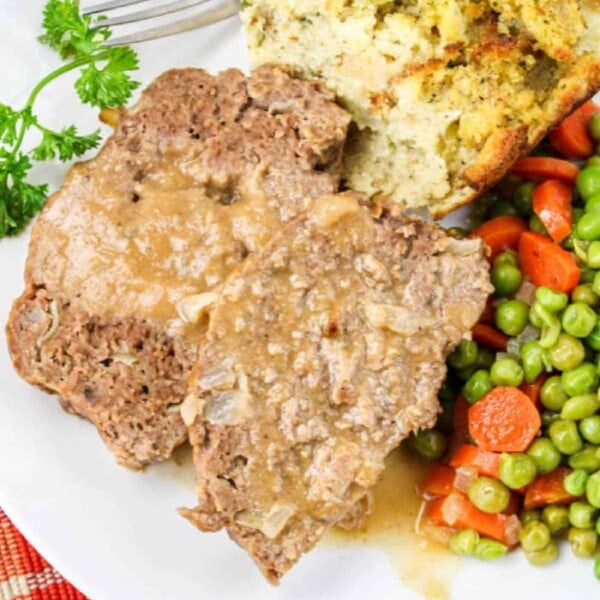 A plate showcasing the savory Onion Lipton Soup meatloaf, accompanied by stuffing and a vibrant mix of peas and carrots on a checkered tablecloth.