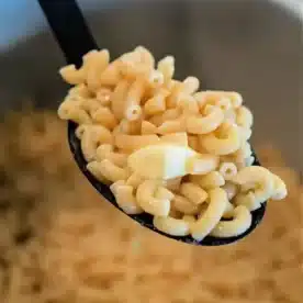 A close-up of a black spoon holding a serving of freshly cooked elbow macaroni, resembling Ninja Foodi butter noodles, over a pot. The light yellow macaroni looks perfectly tender and ready to savor.