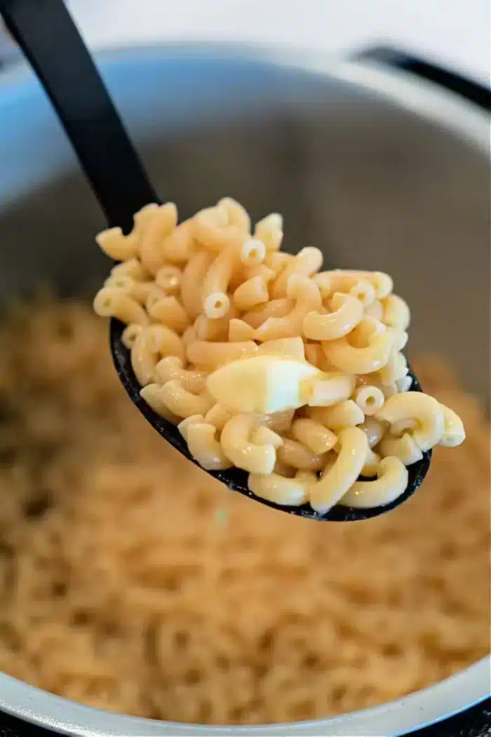 A close-up of a black spoon holding a serving of freshly cooked elbow macaroni, resembling Ninja Foodi butter noodles, over a pot. The light yellow macaroni looks perfectly tender and ready to savor.