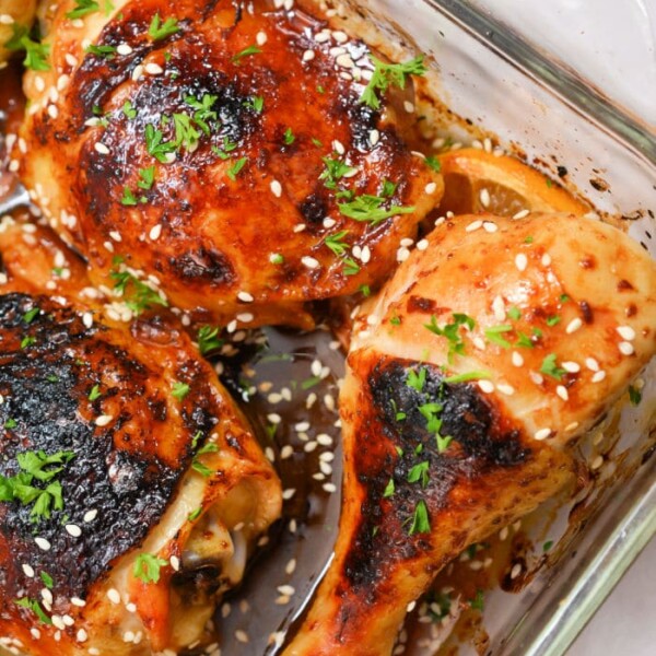 A glass baking dish features Teriyaki chicken drumsticks and thighs, garnished with sesame seeds and chopped herbs. The Cheesecake Factory-inspired chicken pieces are cooked to golden brown perfection with charred areas, and onion slices peek from underneath.