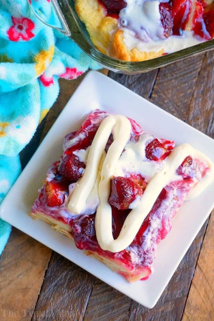 A square piece of strawberry bread pudding topped with fresh strawberries and a white drizzle rests on a white plate, placed on a wooden surface. A blue cloth with a floral pattern is partially visible in the background.