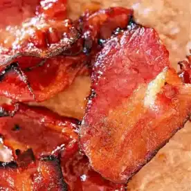 Close-up of crispy, cooked bacon slices on a brown surface. The bacon appears glossy and caramelized, reminiscent of air fryer maple candied bacon.