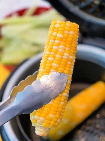 Tongs holding air fryer corn on the cob hover over the basket, showcasing a perfectly cooked ear. Another ear rests inside, ready to savor. Out-of-focus corn husks linger in the background, adding rustic charm to this modern cooking classic.