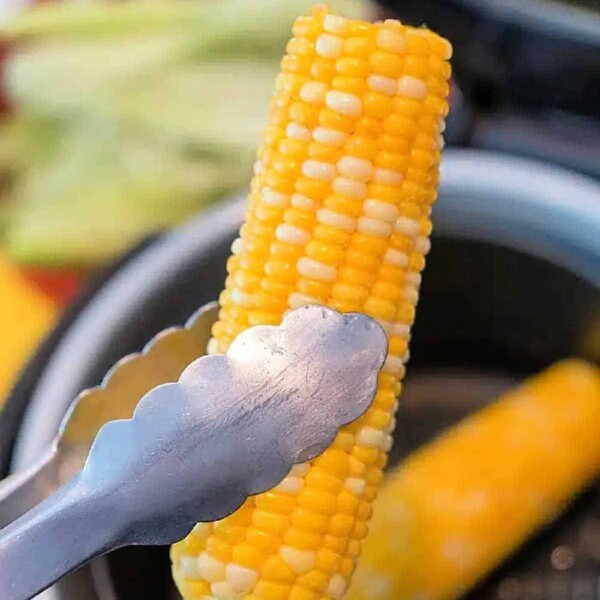 Tongs holding air fryer corn on the cob hover over the basket, showcasing a perfectly cooked ear. Another ear rests inside, ready to savor. Out-of-focus corn husks linger in the background, adding rustic charm to this modern cooking classic.