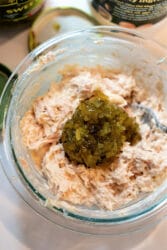 A glass bowl brimming with a creamy blend, perhaps reminiscent of a tuna melt, adorned with chopped pickles. A silver spoon peeks from the side, while jar lids linger in the background.