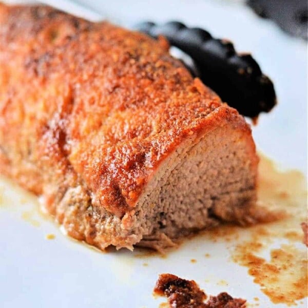 Seasoned pork tenderloin sliced at one end, with flavorful juices on a white surface and tongs in the background—perfect for showcasing how to cook Smithfield pork tenderloin to juicy perfection.