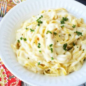 A white bowl filled with creamy Instant Pot fettuccine alfredo, garnished with chopped parsley. The bowl is placed on a colorful patterned tablecloth, with a fork nearby.