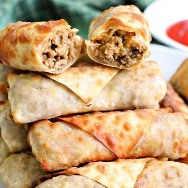 A stack of cooked egg rolls is piled on a plate, one cut in half to reveal the savory filling inside, perfect for using up leftover taco meat. A small dish of red dipping sauce stands ready in the background, set atop a vibrant green cloth.