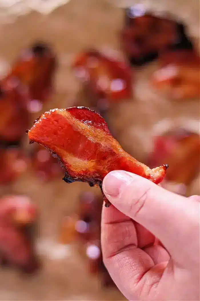 A hand holds a crispy piece of air fryer maple candied bacon with a glossy surface. Other pieces are blurred in the background, scattered on a light-colored surface.