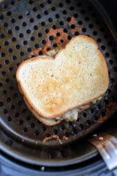 A toasted tuna melt sits inside an air fryer basket. The bread is golden brown, with melted cheese oozing around the edges. The perforated surface below is part of the air fryer.