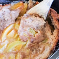 A close-up of a Crockpot cobbler with frozen fruit. A wooden spoon partially lifts the cobbler, revealing juicy sliced peaches and a golden brown, baked surface. The dessert looks moist and freshly cooked.