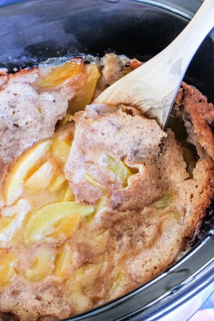 A close-up of Crockpot Cobbler with Frozen Fruit. A wooden spoon scoops out a portion, revealing sliced peaches and a golden-brown, crumbly crust. The dessert appears warm and freshly made.