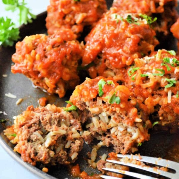 A plate of Instant Pot porcupine meatballs covered in tomato sauce is served. Some are garnished with chopped parsley. A fork rests beside the dish, and one meatball is partially cut open, revealing its savory interior. Parsley adorns the background.