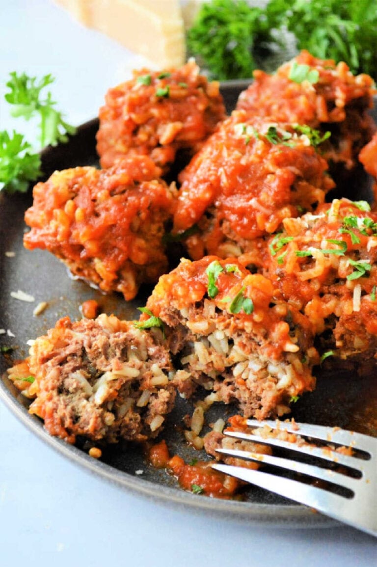 A plate of Instant Pot porcupine meatballs covered in tomato sauce is served. Some are garnished with chopped parsley. A fork rests beside the dish, and one meatball is partially cut open, revealing its savory interior. Parsley adorns the background.