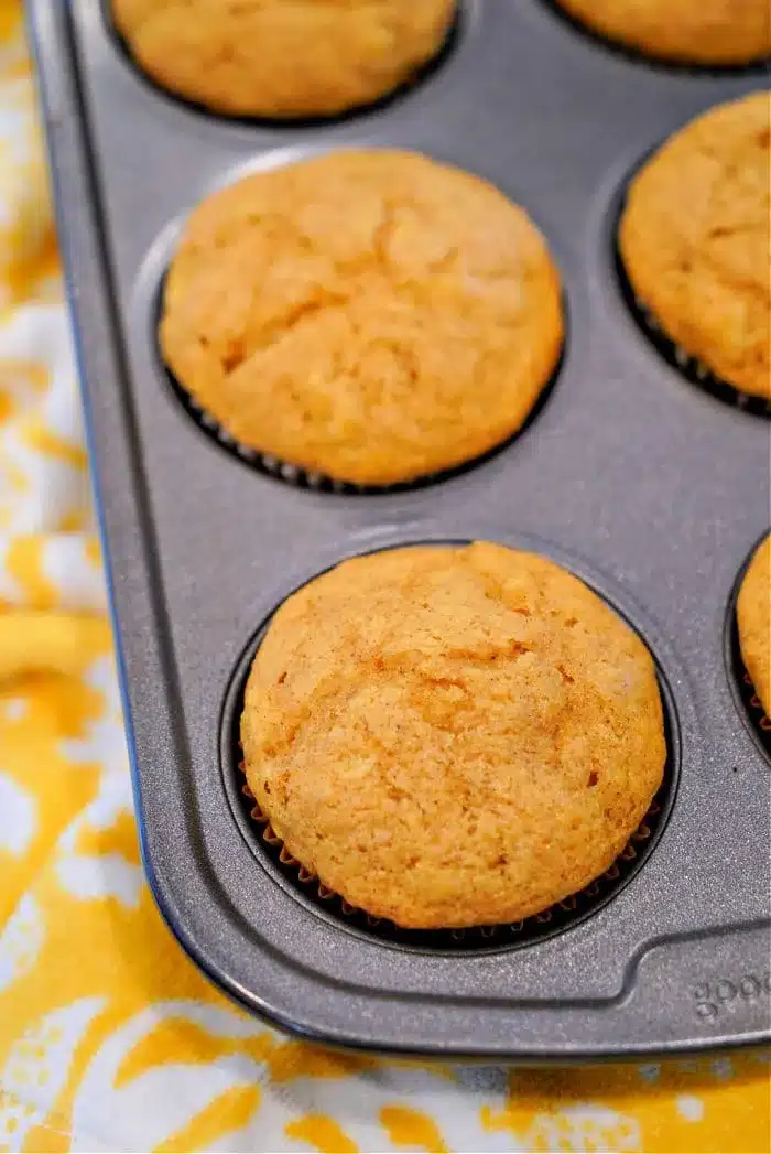 Pumpkin-flavored banana muffins with a golden-brown top are baked in a dark muffin tray, resting on a cheerful yellow and white patterned cloth.