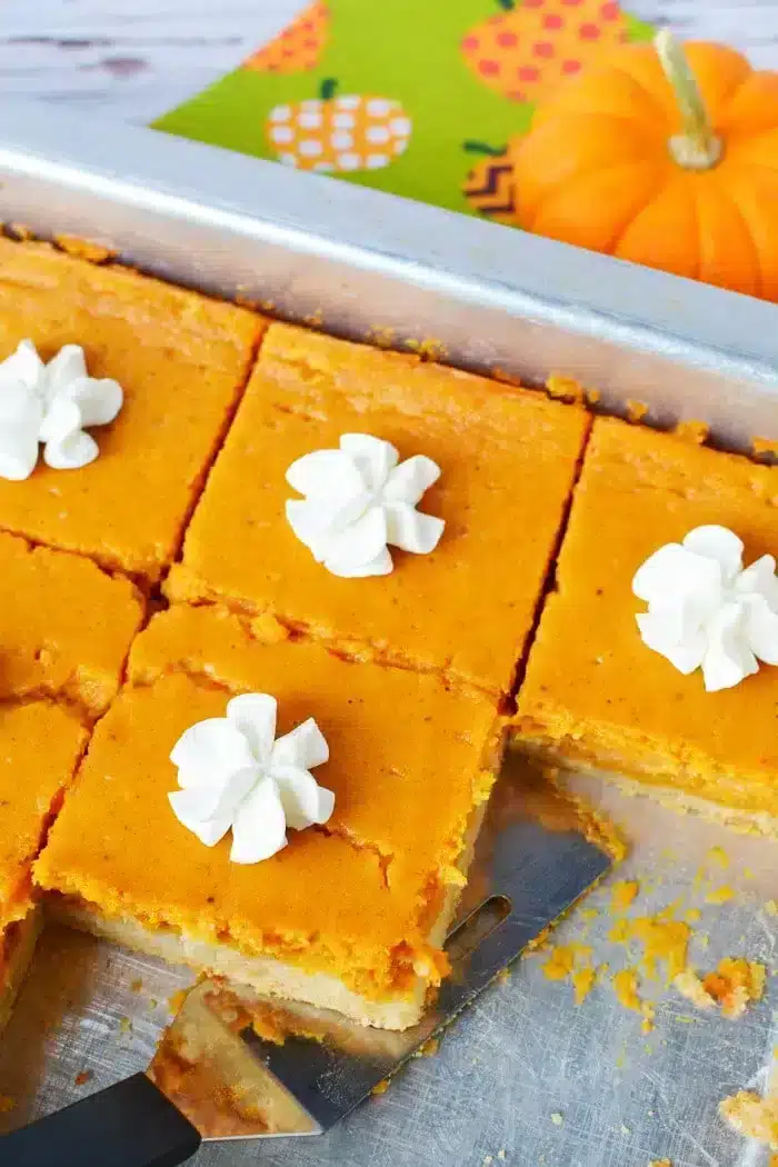 A baking tray contains square pumpkin pie slices, each topped with a dollop of whipped cream. One slice is partially removed with a spatula placed underneath it, almost like cutting into a 1/2 sheet cake. 